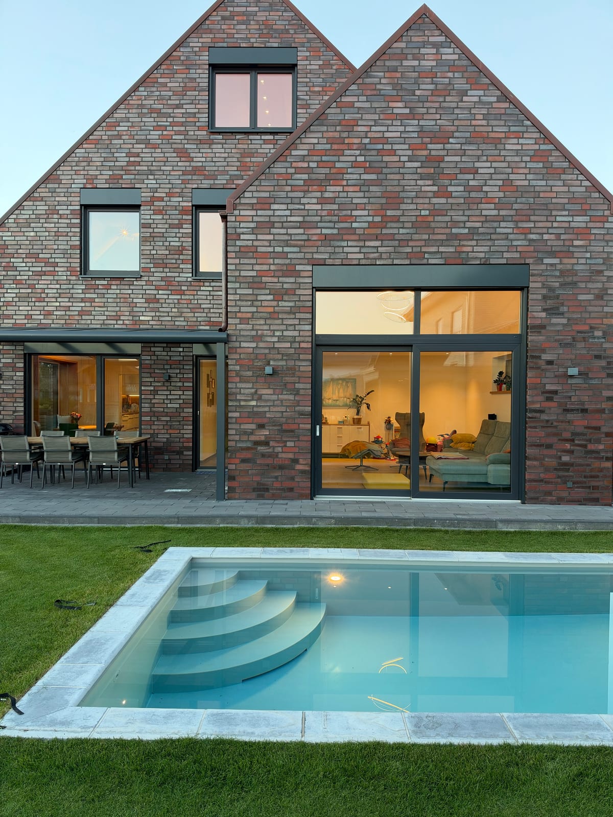 Gartenseite unseres Hauses in der Dämmerung, alle Raffstores oben, Licht im Wohnzimmer, beleuchteter Pool davor