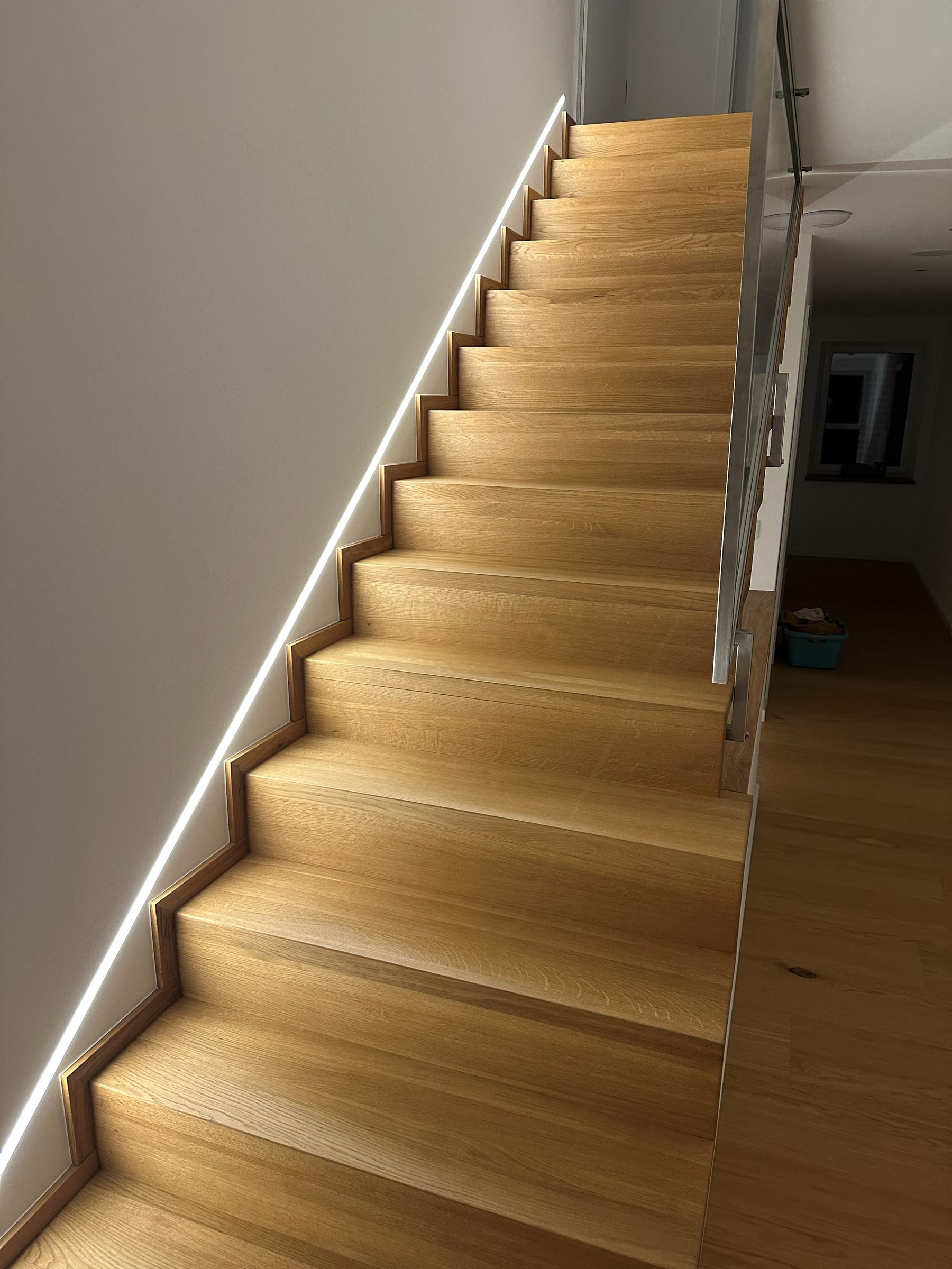 Treppe mit Glasgeländer und aktivierter Akzentbeleuchtung im Abendmodus, Blick vom Obergeschoss ins Dachgeschoss