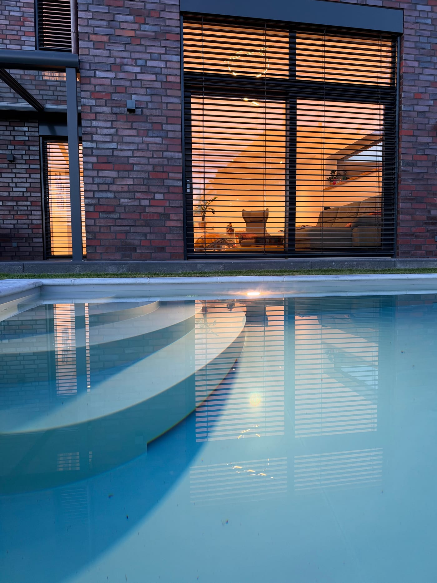 Unser Pool am Abend mit Unterwasserscheinwerfer, im Wasser spiegelt sich das Wohnzimmerfenster mit gewendeten Raffstores